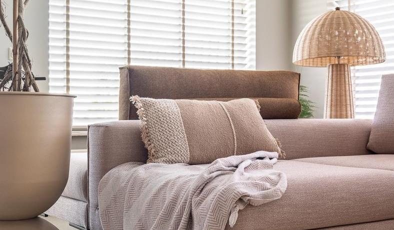 Helles Wohnzimmer mit beigem Sofa, Decke, Kissen, großem Blumentopf und Stehleuchte.