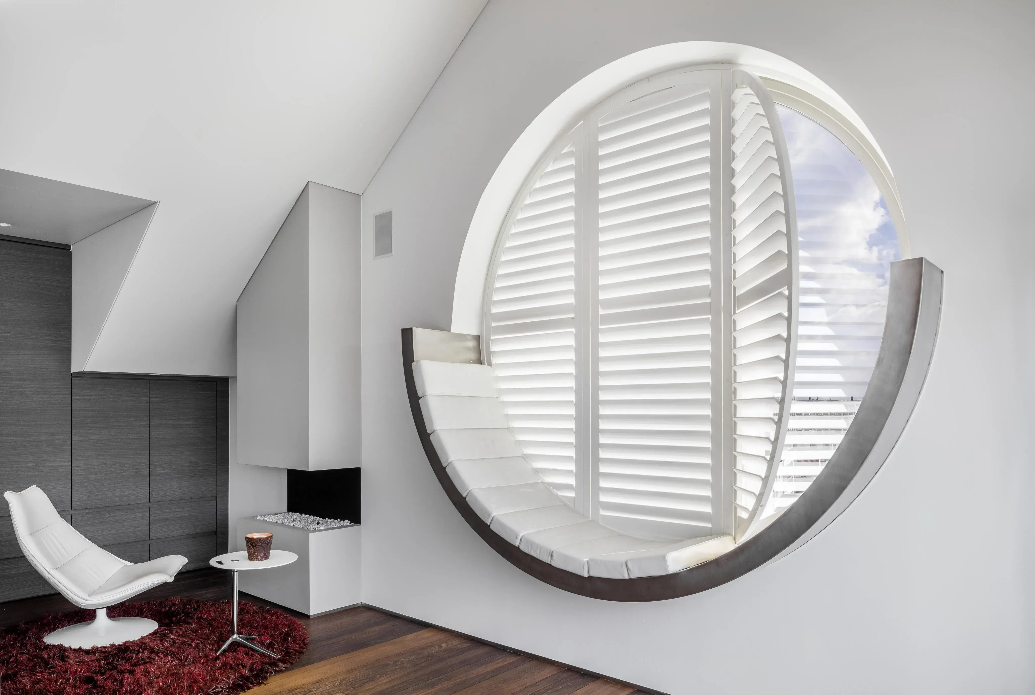 Modernes Wohnzimmer mit rundem Fenster und weißen Lamellen, Sessel und roter Teppich.